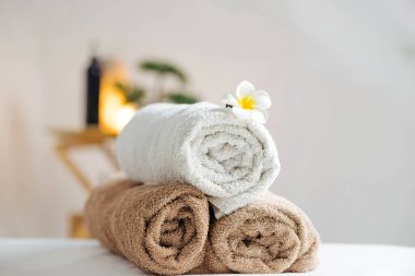 Üzerinde bir plumeria çiçeği olan beyaz ve kahverengi renkli havlular istiflenmiş. Yumuşak ışıklandırmalı huzurlu bir sağlık ortamında bir spa masasına kurulmuş.
