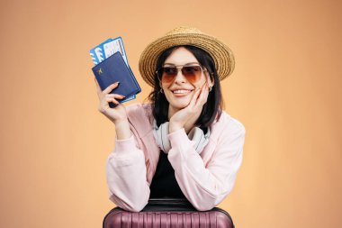 Güneş gözlüklü ve hasır şapkalı genç bir kadın elinde pasaport ve biniş kartı tutarken gülümsüyor, şeftali arkaplanlı mor bir valize yaslanıyor.
