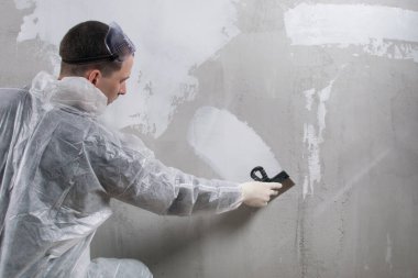 Koruyucu üniformalı bir işçi beton duvara beyaz macun sürer.