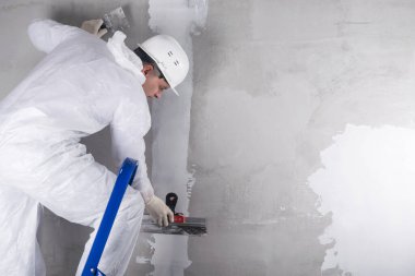 Miğferli ve takım elbiseli bir işçi merdiven üzerinde durur ve karışımı dengelemek için duvara uygular.