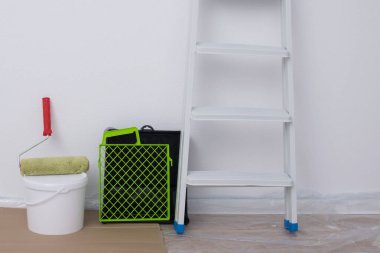 Beyaz bir duvarın arkasında bir merdiven, bir rulo banyo ve bir kova boya var.