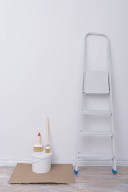 Hazırlanmış yüzeyde, polietilen kaplı bir merdiven, bir kova boya ve bir ressam için fırçalar var.