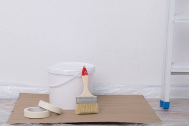Bir kova boya, koli bandı ve fırça, beyaz bir duvarın arka planında.