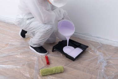 Koruyucu giysiler içindeki bir ressam fırçalara ve silindirlere başvurmak için bir kovadan küvete boya döker.