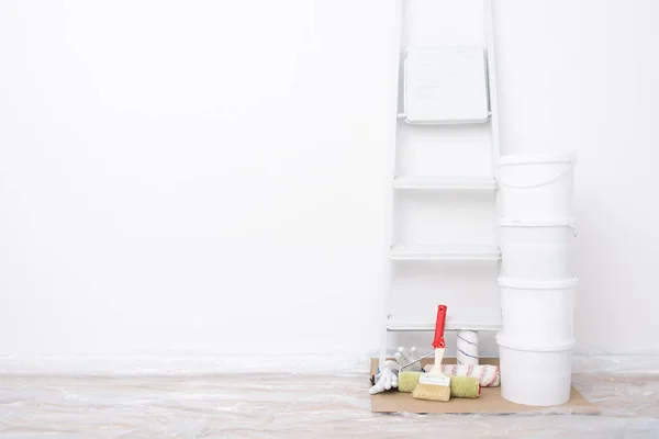 white buckets of paint and a ladder are to the right of the white wall, to the left is space for an inscription