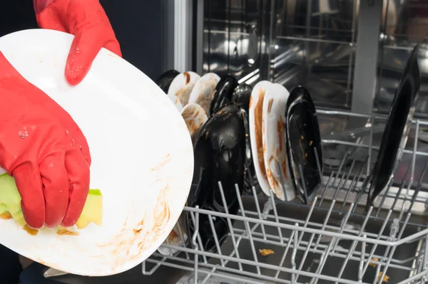 hands in red rubber gloves remove large leftovers from dirty dishes and put them in the dishwasher