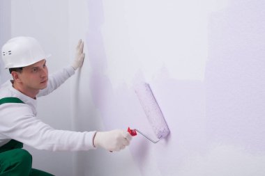 Koruyucu giysili bir işçi, duvarı açık mor boyayla boyuyor, silindirli, yazıtlar için bir yer var.