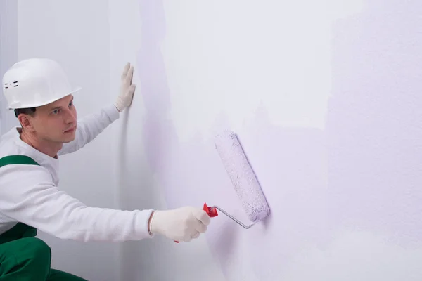 a worker in protective clothing, paints the wall with light purple paint with a roller, there is a place for the inscription