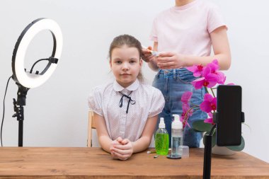 Mola sırasında, yüzük lambasının ışığı altında, blogcu kız, masada, görüntülü konuşma için hazırlanıyor, asistan makyaj yapıyor.