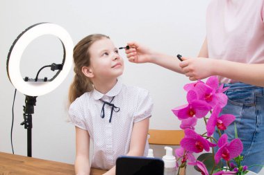 Mola sırasında, yüzük lambasının ışığı altında, blogcu kız, masada, video sohbeti için hazırlanıyor, asistanı kirpiklerine rimel sürüyor.