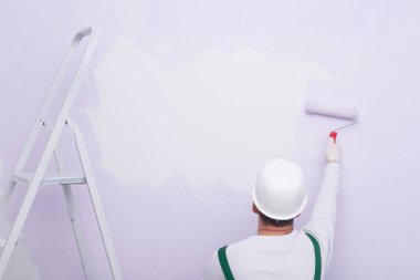 Koruyucu giysili ve kasklı bir inşaat işçisi, duvara silindir ve dikiz aynasıyla boya sürüyor, yazılar için bir yer var.