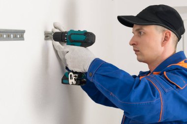 Bir işçi elektrikli tornavida ve yan görüş kullanarak mutfak dolaplarını asmak için kendini tıkırdatan vidaları olan beyaz bir duvara vidaları vidalıyor.