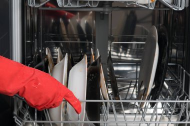 Kırmızı koruyucu eldivenli bir el bulaşık makinesinin ağını kirli tabaklarla doldurur..
