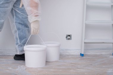 Koruyucu giysili bir adam, beyaz bir duvar ve merdivenin arka planına, yakın plan boya kovaları getiriyor.