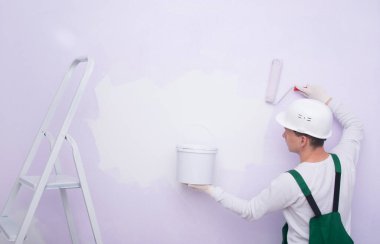 Koruyucu giysili ve kasklı bir inşaat işçisi, rulo boyayla boya sürüyor ve elinde bir kova tutuyor, katlanan merdivenin yanında.