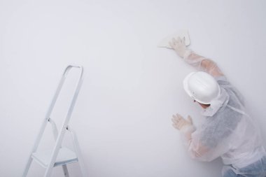 Koruyucu giysili ve başlıklı bir inşaat işçisi, duvar kağıdını plastik bir spatulayla dümdüz eder, duvarlardan ve dikiz aynasından fazla yapıştırıcı çıkarır.