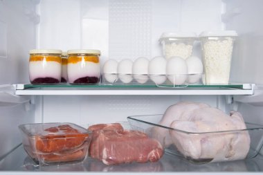 against the background of a white refrigerator, on glass shelves, there are products, whole chicken,fish, meat, eggs, cottage cheese, yogurt clipart