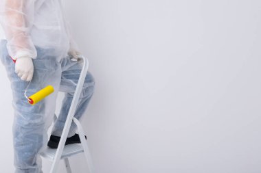 Koruyucu giysiler içindeki bir inşaat işçisi beyaz duvardaki duvar kağıdını özel bir silindirle düzleştirecek. Yazıtlar için bir yer var.