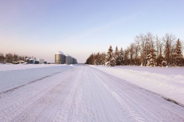 Kırsal çayır yolu kar yağışı sonrası sürüldü, tahıl ambarları ve çalılarla çiftliğe sürüldü, kış manzarası.