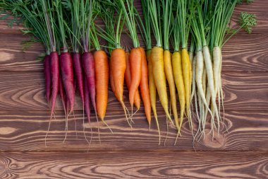 Renkli sarı, kırmızı ve turuncu havuçlar (Gökkuşağı karışımı) ve yeşil yapraklı beyaz (Luna beyaz çeşitliliği) kahverengi ahşap masa üzerindedir. Taze sonbahar mahsulü.