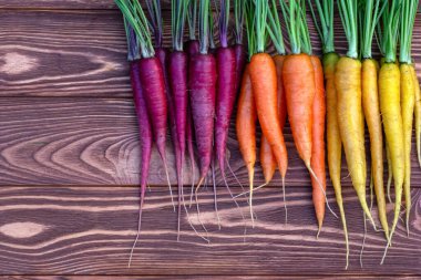 Kahverengi ahşap masanın üzerinde renkli organik sarı, kırmızı ve turuncu havuçlar (gökkuşağı karışımı) ve yeşil yapraklar bulunmaktadır. Taze sonbahar mahsulü.