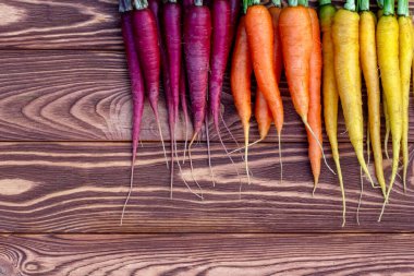 Kahverengi ahşap masanın üzerinde renkli organik sarı, kırmızı ve turuncu havuçlar (gökkuşağı karışımı) ve yeşil yapraklar bulunmaktadır. Taze sonbahar mahsulü.