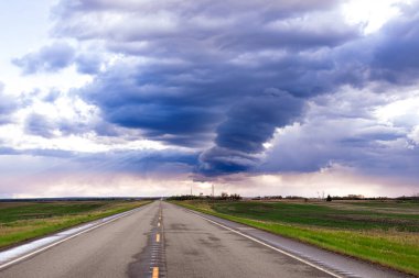 Ovalardaki kırsal alan, tarımsal yeşil tarlalar arasındaki yağmurdan sonra ıslak köy yolu ve yukarıdaki dramatik mavi bulutlu gökyüzü ve güneş ışınları yarılıyor..