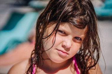 Close-up portrait of a young girl with wet hair in a pink swimsuit, looking intently at the camera under bright sunlight near a swimming pool.