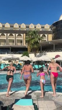 Kemer, Turkey - September 21, 2025 - Four people, including a child, jump into a hotel swimming pool on a sunny day. The backdrop features umbrellas, sunbeds, and a modern hotel building.