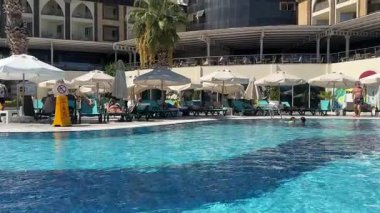 Kemer, Turkey - September 21, 2025 - Smooth sliding shot of a sunny hotel poolside with people relaxing on loungers under umbrellas, palm trees, and a modern resort building in the background.