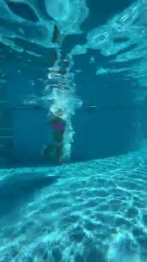 A young woman swims elegantly underwater in a pool, showcasing her fluid motion as the camera follows her, highlighting the tranquil blue water and reflections