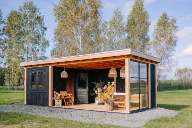 Modern black wooden cabin with a covered terrace, cozy seating, rattan lights, and floral decorations, set in a green field surrounded by autumn trees.