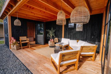 Modern wooden terrace with white cushioned seating, wicker pendant lights, black walls, and cozy decor. Door open to small wooden interior space.