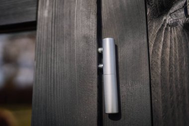 Close-up of a silver metal door hinge mounted on a black painted wooden frame, showcasing modern minimalist hardware detail in natural lighting.