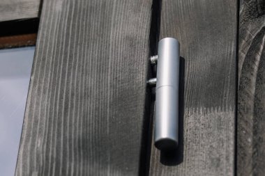 Close-up of a metallic silver hinge mounted on a black wooden surface, showing detailed texture of wood grain and modern hardware under natural light.