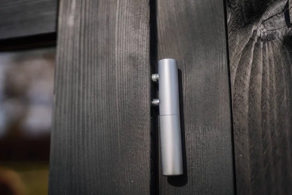 Close-up of a silver metal door hinge mounted on a black painted wooden frame, showcasing modern minimalist hardware detail in natural lighting.