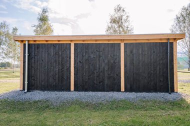 Rear view of a modern wooden cabin with vertical charred black timber cladding, light wood support beams, and gravel foundation, surrounded by grassy landscape.