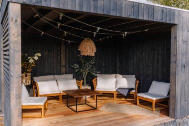 Modern outdoor gazebo with cozy white-cushioned wooden seating, string lights, rattan lamp, potted plants, and dark wood paneling for a stylish retreat.