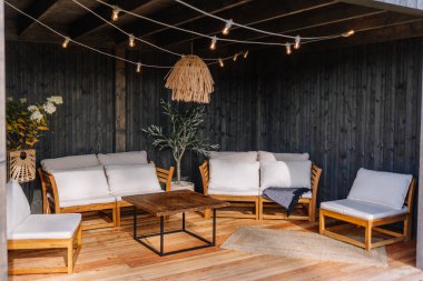 Cozy outdoor lounge with wooden furniture, white cushions, string lights, rattan pendant lamp, and potted plants in a black wood-paneled garden gazebo.