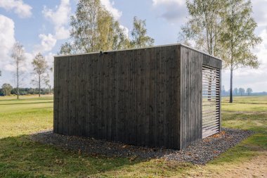 A modern dark wooden gazebo with vertical cladding and slatted wall, situated in a grassy open field surrounded by trees under a bright cloudy sky.
