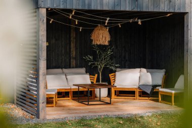 Cozy modern wooden gazebo with light string bulbs, cushioned seating, a wooden coffee table, and decorative plants, creating a stylish outdoor lounge space.