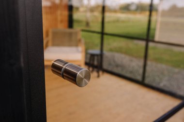 Modern stainless steel door handle on a glass door, leading to a bright room with wooden flooring and outdoor view through large glass windows.
