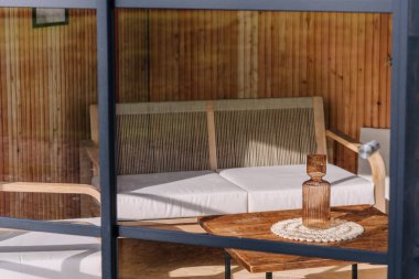 Close-up of a cozy modern lounge seen through glass, featuring a wooden sofa with white cushions and a rustic table with a decorative glass bottle in sunlight.