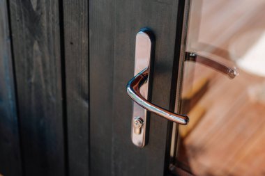 Close-up of a shiny chrome door handle on a black wooden and glass door, reflecting warm sunlight and natural wood tones from the surrounding interior.