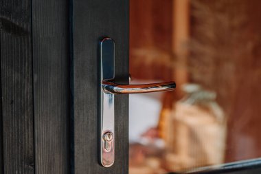 Modern chrome door handle on a dark wooden door with glass panel reflecting warm interior decor, natural wood textures, and soft lighting.