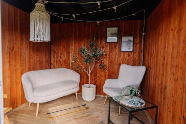 Stylish tiny house interior with wooden walls, a cozy white sofa and armchair, a potted plant, macrame lamp, rug, framed art, and a decorative coffee table.