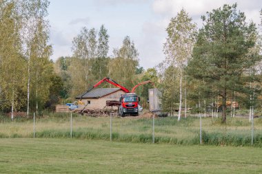 Sigulda, Latvia - October 7, 2025 - A red MAN truck with a crane arm operates at a rural construction or demolition site near a brick building, surrounded by birch trees and green fields.