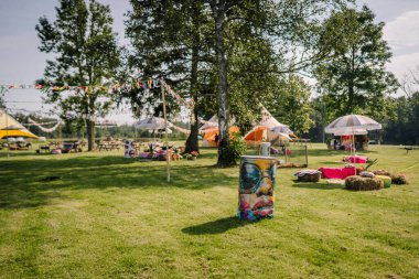Saldus, Latvia - July 19, 2025 - Colorful hippie festival setup in a grassy park with decorated tents, painted barrels, hay seating, parasols, and ribbon garlands hanging between trees.