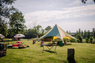Saldus, Latvia - July 19, 2025 - Spacious outdoor setting of a hippie festival with a large canopy tent, hammock, hay bale seating, colorful decorations, and trees under a bright sky.