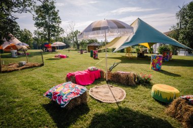 Saldus, Latvia - July 19, 2025 - Colorful bohemian seating area at a summer hippie festival, featuring hay bales, umbrellas, rugs, and vibrant decorations on a sunny open lawn.